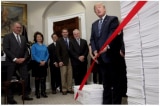 Tổng thống Donald Trump cắt tấm băng đỏ giữa hai chồng giấy tờ, tượng trưng cho các quy định của chính phủ trong những năm 1960, và các quy định của ngày hôm nay, hôm 14/12/2017